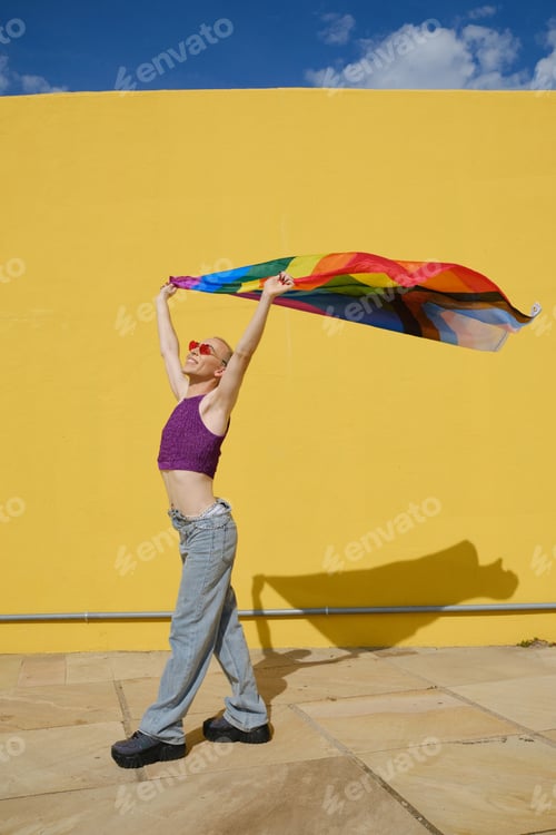 Preview: Happy young non-binary person holding and waving a rainbow flag outdoors.
