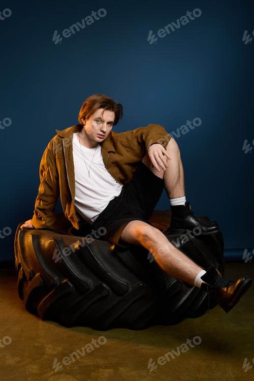 Preview: Fashionable young man poses confidently on a large tire in a creative studio setting