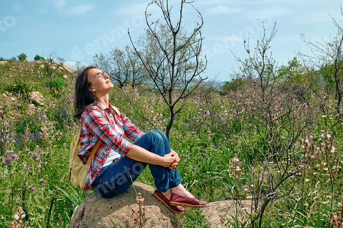 Preview: Happy carefree woman sitting on big stone in a spring meadow with beautiful field flowers.
