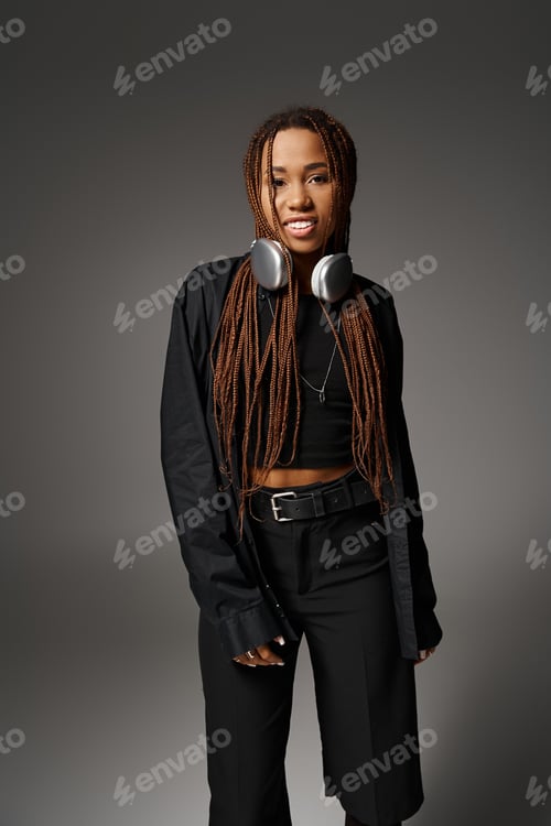 Preview: happy african american woman with dreadlocks standing in headphones and looking at camera on gray