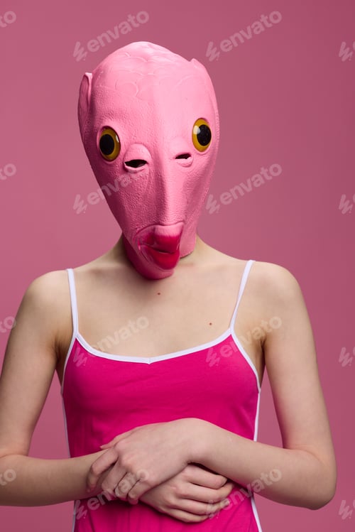 Preview: A woman wearing a pink fish head mask on Halloween stands and looks at the camera against a pink