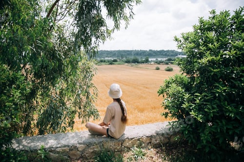 Preview: a teenage girl with a braid in a t-short and hat sits outdoor and looks at the wheat field