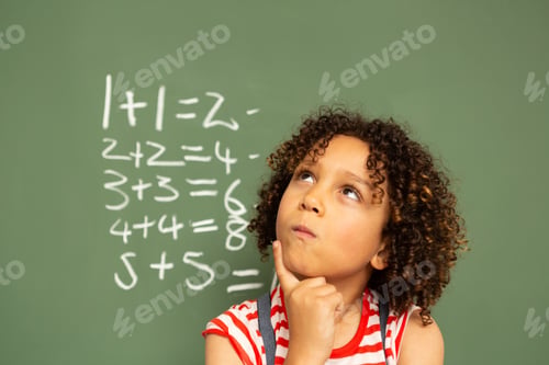 Preview: Schoolgirl standing against green board with one finger on the chin in a classroom at school
