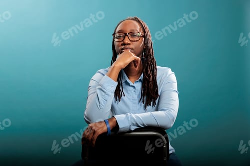 Preview: Serious confident woman sitting on chair while looking at camera on blue background.