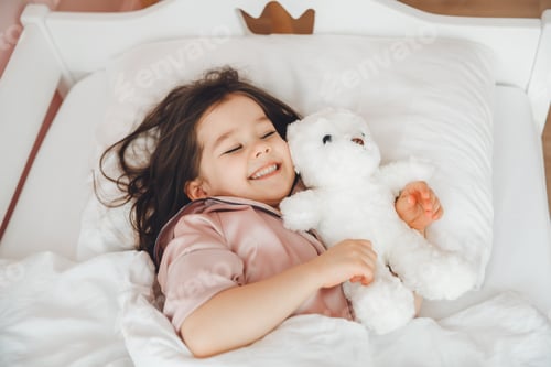 Preview: a little dark-haired girl is sleeping in bed, hugging a teddy bear under a white blanket.the child