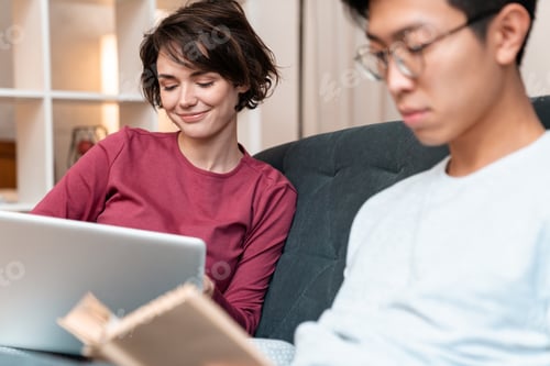 Preview: Photo of pleased multinational couple reading book and using laptop