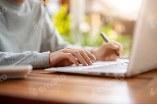 Preview: Close up of a hand holding a pen and other hand touching on laptop while sitting at wooden table.
