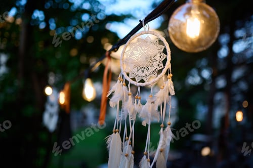Preview: Dreamcatcher with soft feathers hanging under glowing lights in the evening, creating a warm and