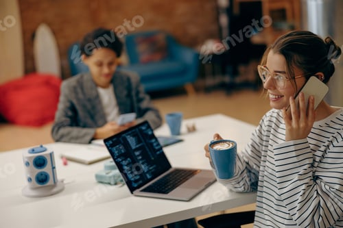 Preview: Female programmer is talking phone while working on laptop and drinking coffee sitting at coworking