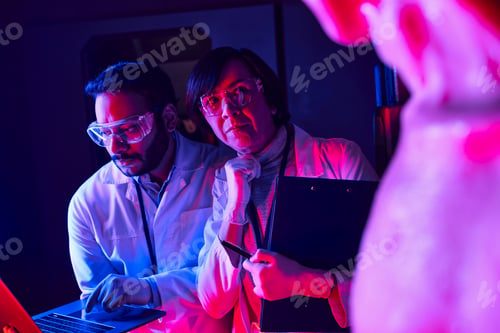 Preview: woman scientist looking at alien near indian colleague using laptop in futuristic science center