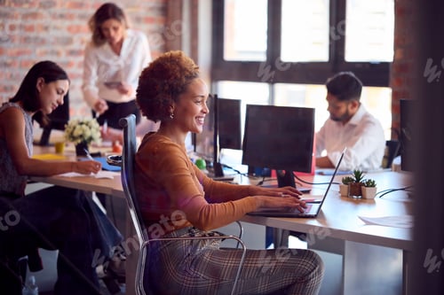 Preview: Businesswoman Working On Laptop At Desk In Busy Multi-Cultural Office