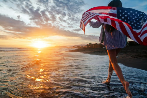 Preview: Beautiful patriotic young woman with the American flag held in her standing in the summer sunset