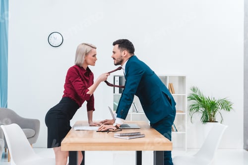 Preview: side view of young businesswoman holding necktie of handsome business man and flirting in office