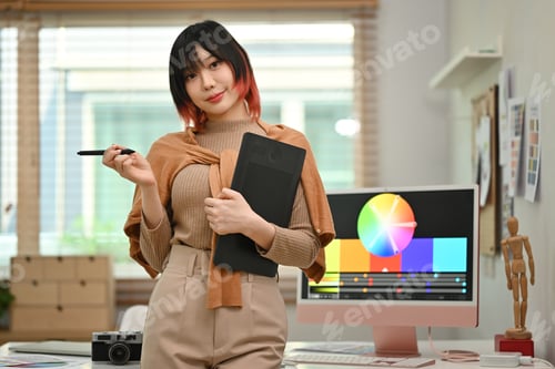 Preview: Stylish young woman holding graphic tablet standing at working desk with color swatch.