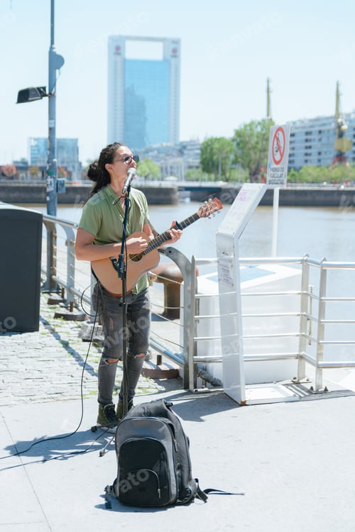 Vista previa: imagen vertical de un joven músico callejero de pelo largo tocando la guitarra y cantando en Puerto Madero, Argentina