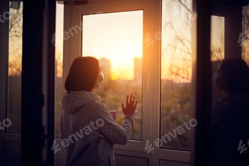 Preview: Woman near window at sunset in isolation at home for virus outbreak. Stay home concept