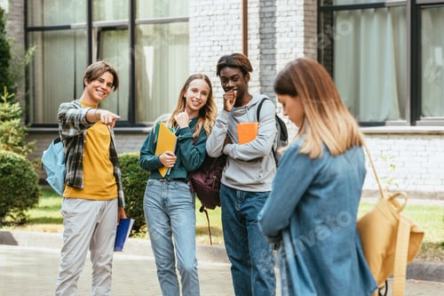 Preview: Selective focus of smiling multicultural teenagers with backpacks pointing at sad girl outdoors