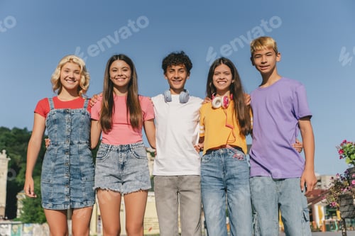 Preview: Group of smiling friends, multiracial teenagers wearing colorful t shirts hugging standing outdoors