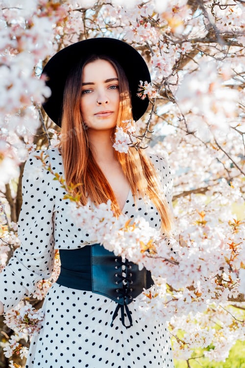 Preview: Young brunette woman in hat and dress near a white blooming tree. Generation Z girl