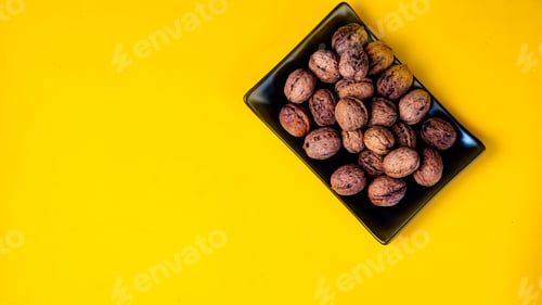Preview: Black plain dish full of nuts on a yellow background.