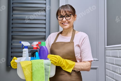 Preview: Middle-aged woman in gloves apron with basin of detergents in bathroom