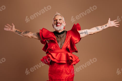 Preview: Joyful gay man with crown on head wearing beautiful red dress and gesturing against brown background