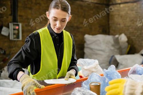 Preview: Young female sorter in protective vest and gloves working with trash on conveyor in blurred waste