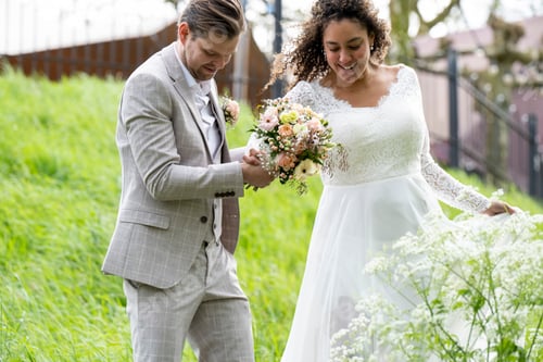 Preview: Bride and groom happily admiring a bouquet together in a lush green setting.