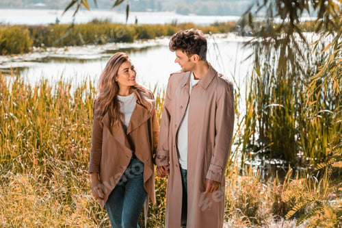 Preview: pleased woman and man in trench coats holding hands near lake in autumnal park