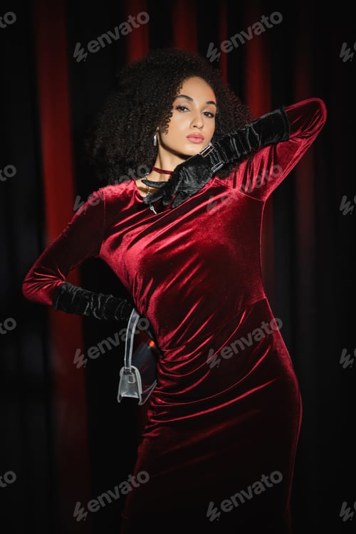 Preview: Trendy african american model in dress holding handbag and standing on burgundy drapery background