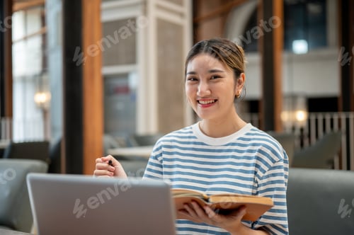 Preview: Smiling asian young woman holding pen and book as sit on sofa in front of a laptop on table in cafe.