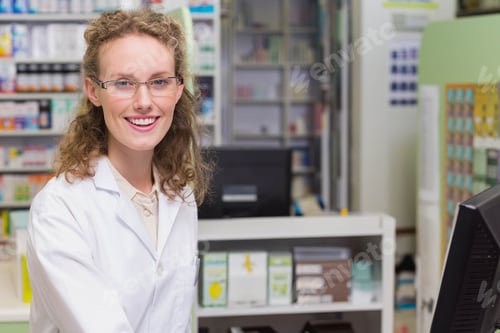 Preview: Smiling female pharmacist in lab coat standing behind pharmacy counter using computer and register