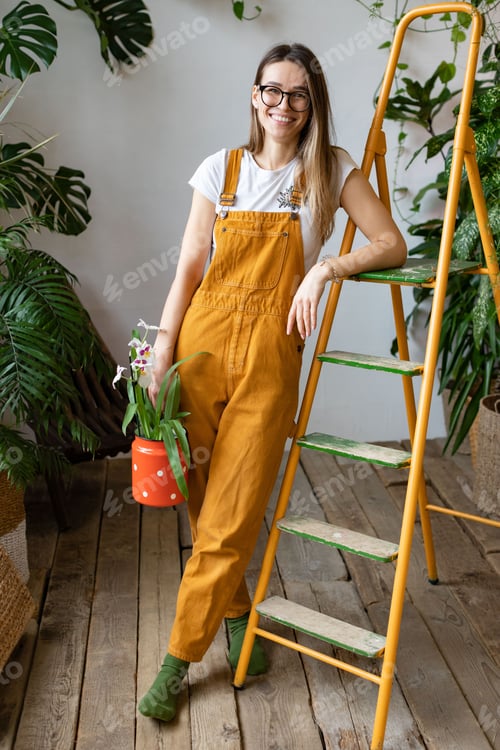 Preview: Woman gardener holding orchid in red milk can, smiling. Home gardening, love of houseplants, hobby.
