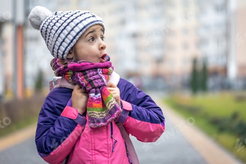 Preview: Little girl with a backpack, in a jacket and a hat near the school, copy space.