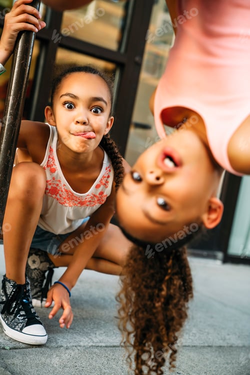 Preview: Happy, smiling girls hanging from bike rack bar on sidewalk