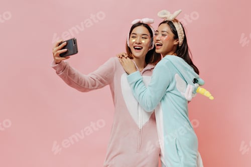 Preview: Cheerful brunette women in kigurumis and headbands smile sincerely and take selfie on pink isolated
