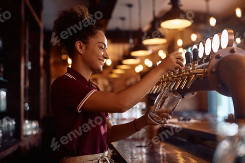 Preview: Happy waitress pouring beer from beer tap at bar counter.