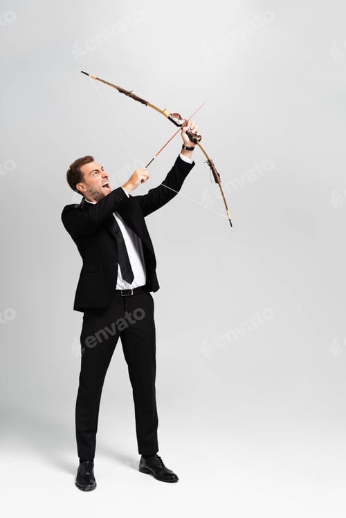 Preview: angry businessman in suit holding bow and shooting with arrow on grey background