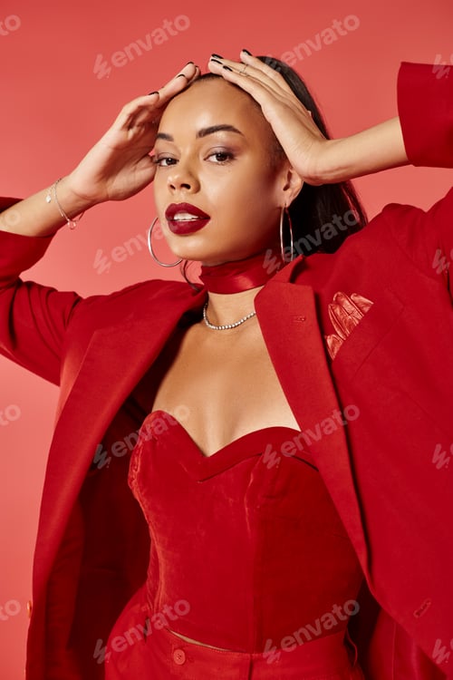 Preview: elegant african american woman with ponytail posing in suit and looking at camera on red backdrop