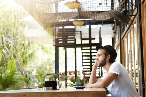 Preview: Stylish Caucasian bearded student in headwear drinking cappuccino alone at modern outdoor coffee sho