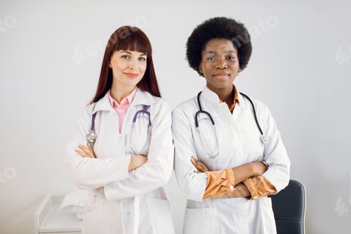 Preview: Two smiling happy African and Caucasian female doctors colleagues, wearing white coats, standing