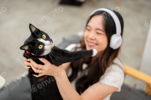Preview: Asian woman listening to music in headphone while holding playing with black cat on living room sofa