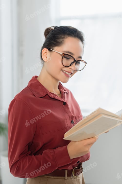 Preview: young teacher in eyeglasses smiling while reading book