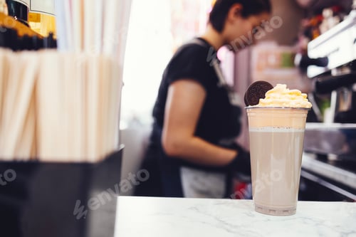 Preview: Young brunette female barista in black shirt, apron and black gloves decorates milk cocktail