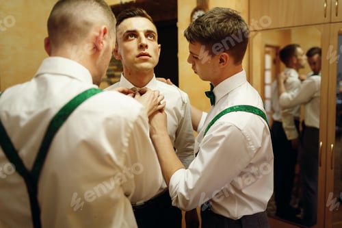 Preview: Handsome groomsman friends helping groom put on stylish white shirt