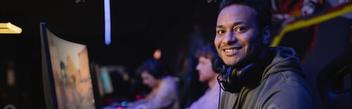 Preview: Positive indian gamer with headphones looking at camera in cyber club, banner