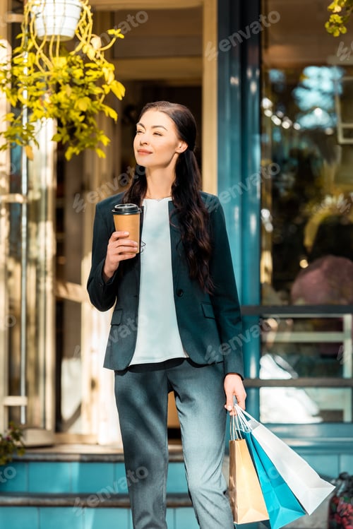 Preview: cheerful beautiful woman standing with shopping bags and disposable coffee cup on street near cafe