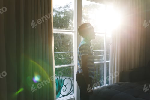 Preview: African american boy thinking and standing at window in sunny living room