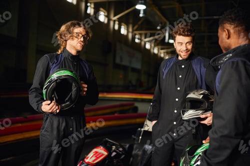 Preview: positive interracial men chatting inside of kart circuit, three kart racers holding helmets