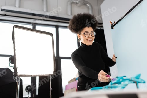 Preview: cheerful african american content maker in eyeglasses looking at camera near reflector and shooting